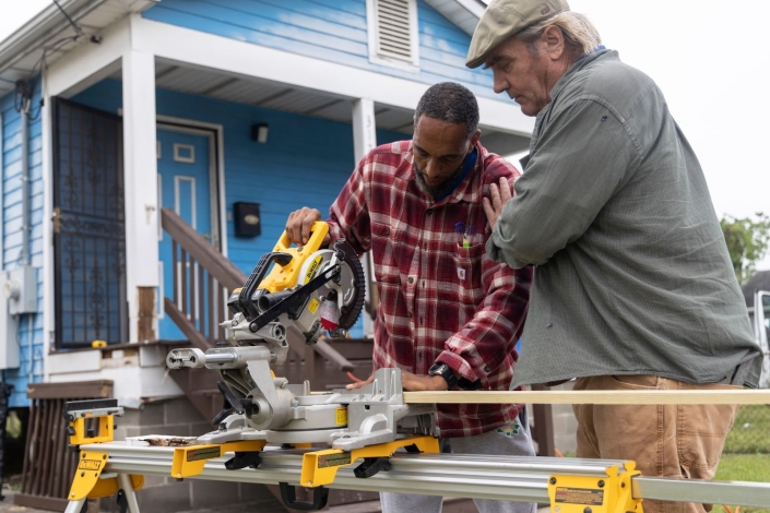 a man uses a circular saw at a job site while his mentor stands over him with his hand on his shoulder in support