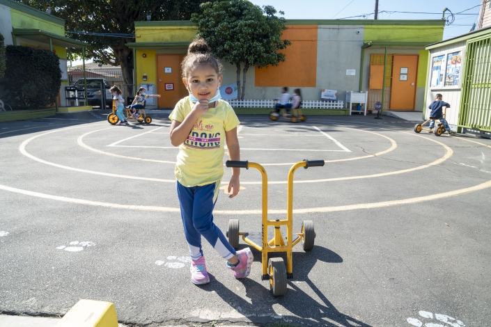 a young girls in a yellow t shirt, blue pants and bright pink shoes stands next to a yellow scooter on a playground to pose for a photo as children play behind her on scooters and push bikes